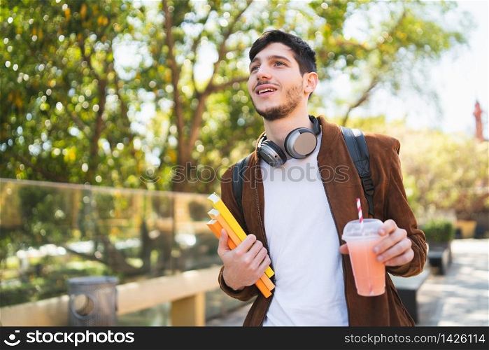 Portrait of young university student walking on the street and holding his books while drinking fresh fruit juice outdoors in the street. Urban and lifestyle concept.