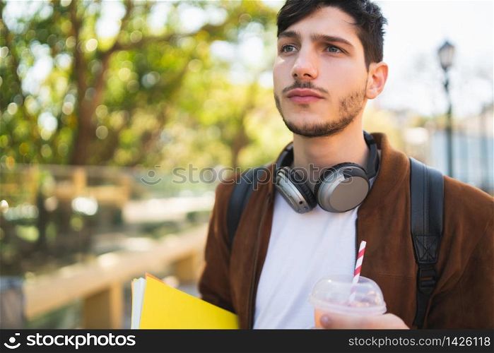 Portrait of young university student walking on the street and holding his books while drinking fresh fruit juice outdoors in the street. Urban and lifestyle concept.