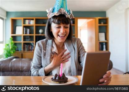 Portrait of young woman celebrating her birthday on a video call with digital tablet and a cake at home. New normal lifestyle concept.. Woman celebrating her birthday on a video call.