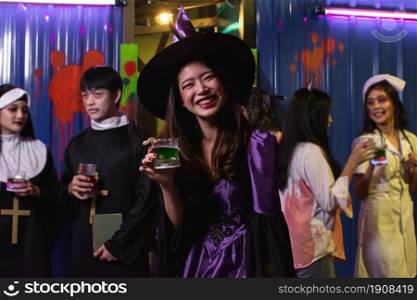Portrait of young woman dressing halloween costume, enjoying and dancing in party while drinking and holding a glass of cocktail with blur background of people