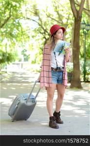 Portrait of young woman holding a map and looking for directions while carrying suitcase outdoors in the street. Travel concept.