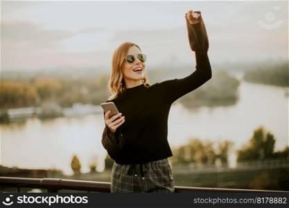 Portrait of young woman holding mobile phone while standing on walkway by the river