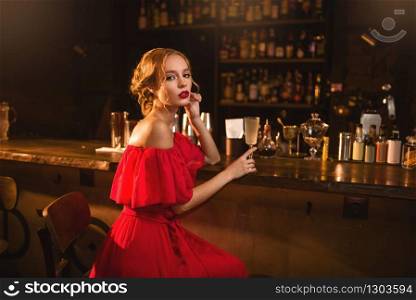 Portrait of young woman in red dress sitting at the bar counter. Beautiful lady with cocktail in hand in club