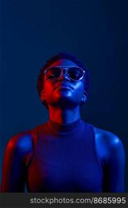 Portrait of young woman in sunglasses looking straight ahead studio shot over dark background with neon light. Woman in sunglasses looking up studio shot over dark