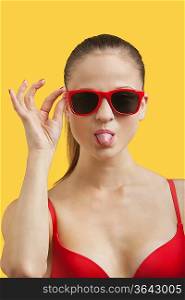 Portrait of young woman in sunglasses sticking out tongue over yellow background