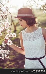 Portrait of young woman in the flowered field in the spring time. Almond flowers blossoms. Girl wearing white dress and sun hat