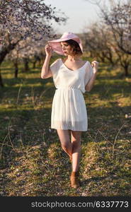 Portrait of young woman in the flowered field in the spring time. Almond flowers blossoms. Girl wearing white dress and pink sun hat