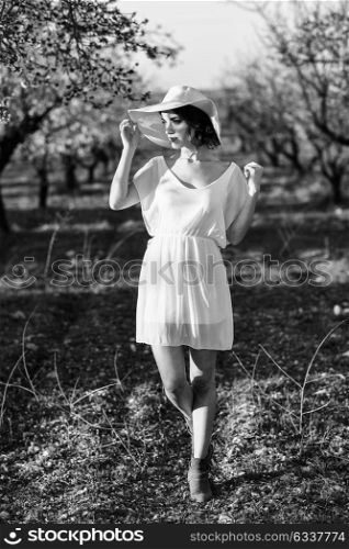 Portrait of young woman in the flowered field in the spring time. Almond flowers blossoms. Girl wearing white dress and pink sun hat