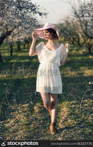 Portrait of young woman in the flowered field in the spring time. Almond flowers blossoms. Girl wearing white dress and pink sun hat