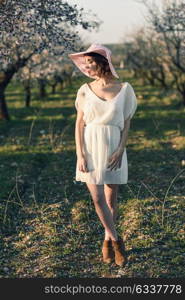 Portrait of young woman in the flowered field in the spring time. Almond flowers blossoms. Girl wearing white dress and pink sun hat