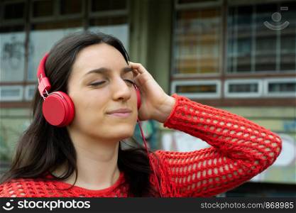 Portrait of young woman listening to music with red headphones. Outdoors.