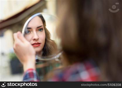 Portrait of young woman looking at herself in a little mirror in urban background.
