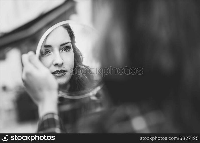Portrait of young woman looking at herself in a little mirror in urban background. Black and white photograph.