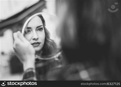 Portrait of young woman looking at herself in a little mirror in urban background. Black and white photograph.