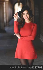 Portrait of young woman, model of fashion, smiling in urban background wearing red dress and hat