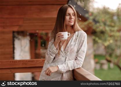 portrait of young woman on the balcony holding a cup of coffee or tea in the morning. She in hotel room looking at the nature in summer. Girl is dressed in stylish nightwear. Relax time