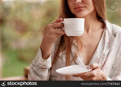 portrait of young woman on the balcony holding a cup of coffee or tea in the morning. She in hotel room looking at the nature in summer. Girl is dressed in stylish nightwear. Relax time