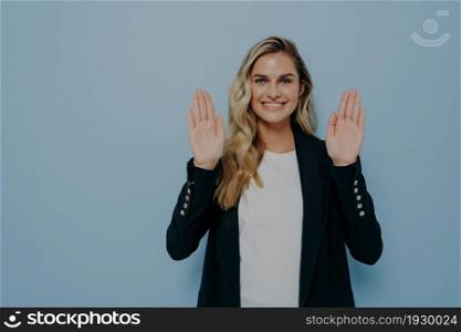 Portrait of young woman refusing offer in polite manner smiling friendly with positive look as waving hands in stop gesture being not interested isolated over blue studio background with copy space. Woman refusing in polite manner smiling friendly with positive look as waving hands in stop gesture
