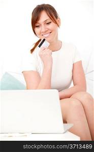 Portrait of young woman sitting on sofa using laptop with card in her hand and giving you an attractive smile