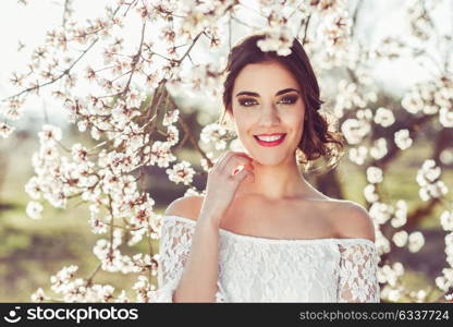 Portrait of young woman smiling in the flowered garden in the spring time. Almond flowers blossoms. Girl dressed in white like a bride.