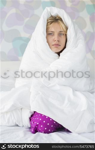 Portrait of young woman suffering from fever while wrapped in quilt in bed