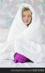 Portrait of young woman suffering from fever while wrapped in quilt in bed