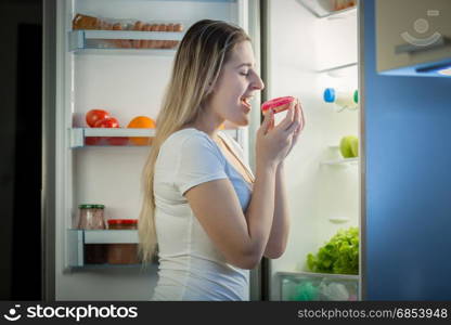 Portrait of young woman taking donut from refrigerator at night. Concept of unhealthy eating