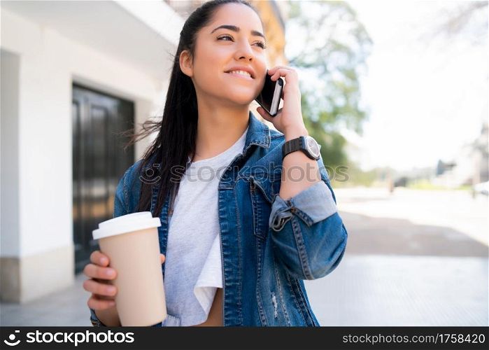 Portrait of young woman talking on the phone and holding a cup of coffee while standing outdoors on the street. Urban and communication concept.