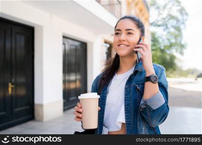 Portrait of young woman talking on the phone and holding a cup of coffee while standing outdoors on the street. Urban and communication concept.