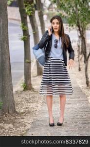 Portrait of young woman talking with smartphone in urban background wearing casual clothes. Girl wearing striped skirt, sweater and leather jacket