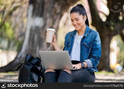 Portrait of young woman using her laptop and drinking coffee while sitting outdoors. Urban concept.