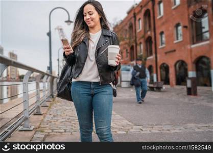 Portrait of young woman using her mobile phone while walking with cup of coffee. Urban and communication concept.