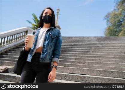 Portrait of young woman wearing face mask and holding a cup of coffee while standing outdoors. Urban concept. New normal lifestyle concept.