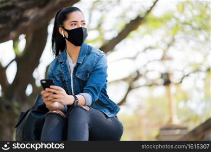 Portrait of young woman wearing face mask and using her mobile phone while sitting on stairs outdoors. New normal lifestyle concept. Urban concept.