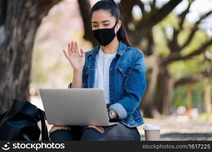 Portrait of young woman wearing face mask on a video call with laptop while sitting outdoors. Urban concept. New normal lifestyle concept.