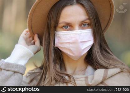 Portrait of young woman wearing hat and protective face medical mask standing in park on sunny day. Virus spread flu prevention quarantine. Portrait of young woman wearing hat and protective face medical mask standing in park on sunny day. Virus spread flu prevention quarantine.