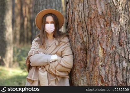 Portrait of young woman wearing hat and protective face medical mask standing in park on sunny day. Virus spread flu prevention quarantine. Portrait of young woman wearing hat and protective face medical mask standing in park on sunny day. Virus spread flu prevention quarantine.