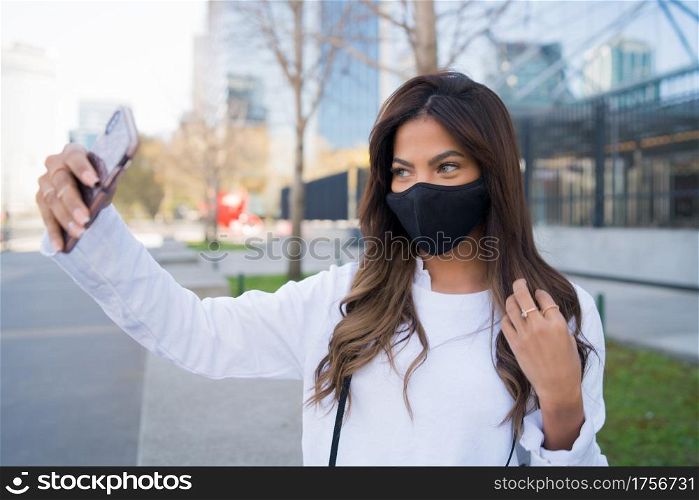 Portrait of young woman wearing protective mask and taking selfies with her mophile phone while standing outdoors. Urban concept. New normal lifestyle concept.