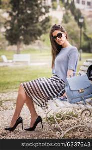 Portrait of young woman with beautiful legs in urban park wearing casual clothes. Girl wearing striped skirt, sweater, sunglasses and high heels