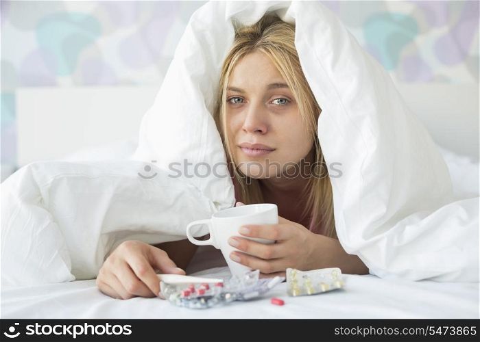 Portrait of young woman with coffee mug and medicines suffering from fever while covered in quilt on bed