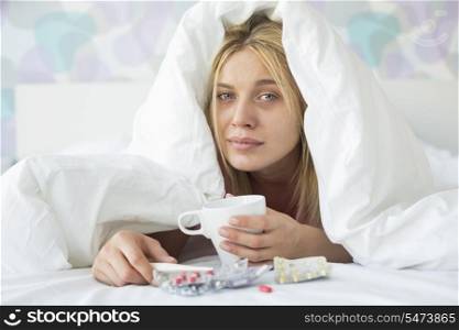Portrait of young woman with coffee mug and medicines suffering from fever while covered in quilt on bed