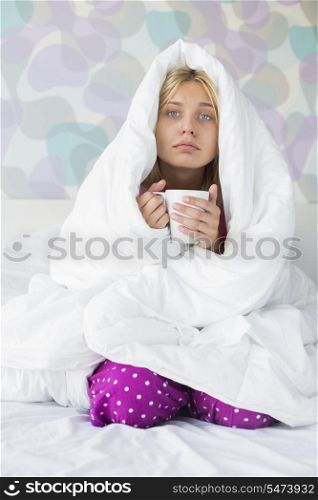 Portrait of young woman with coffee mug suffering from fever while wrapped in quilt on bed