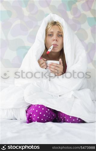 Portrait of young woman with coffee mug taking temperature while wrapped in quilt on bed