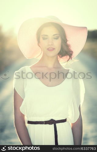 Portrait of young woman with eyes closed in a rural road in the spring time. Girl wearing white dress, brown belt and pink sun hat. Backlight photography with the Sun in the back of the woman.