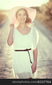 Portrait of young woman with eyes closed in a rural road in the spring time. Girl wearing white dress, brown belt and pink sun hat. Backlight photography with the Sun in the back of the woman.