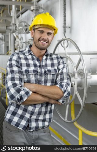 Portrait of young worker with arms crossed leaning on large valve in industry