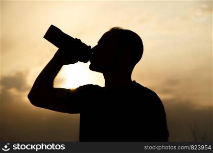 portrait ,outline of a sporty young Caucasian guy in a black t-shirt and black shorts drinking water from a bottle at sunset