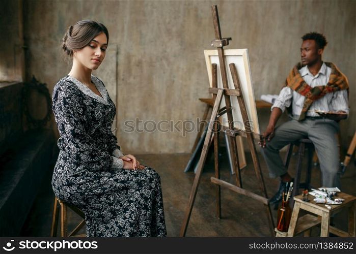 Portrait painter with palette and brush draws female model. Man sitting at the easel, art studio interior on background. Painter with palette and brush draws female model