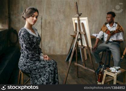 Portrait painter with palette and brush draws female model. Man sitting at the easel, art studio interior on background. Painter with palette and brush draws female model