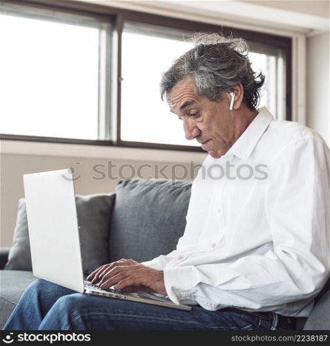 portrait senior man sitting sofa wearing bluetooth earphone using laptop